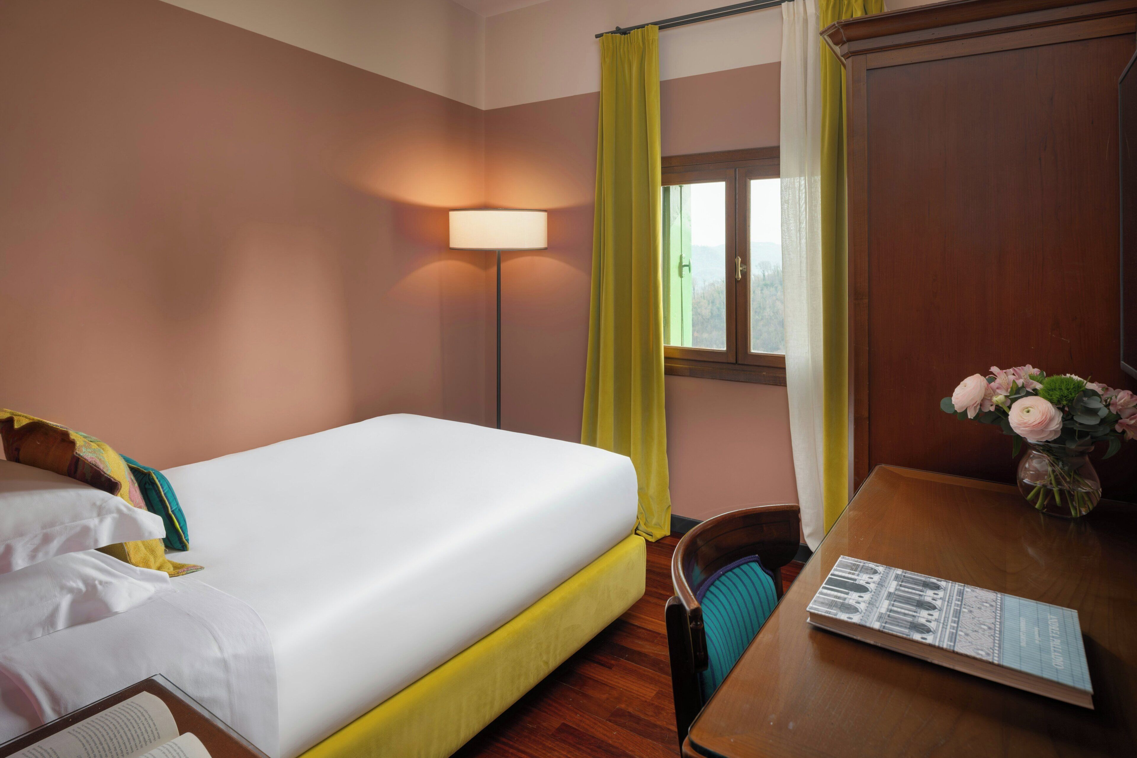 Une chambre d’hôtel avec un lit, un bureau doté d’une lampe, une chaise, une fenêtre avec des rideaux et un vase avec des fleurs.