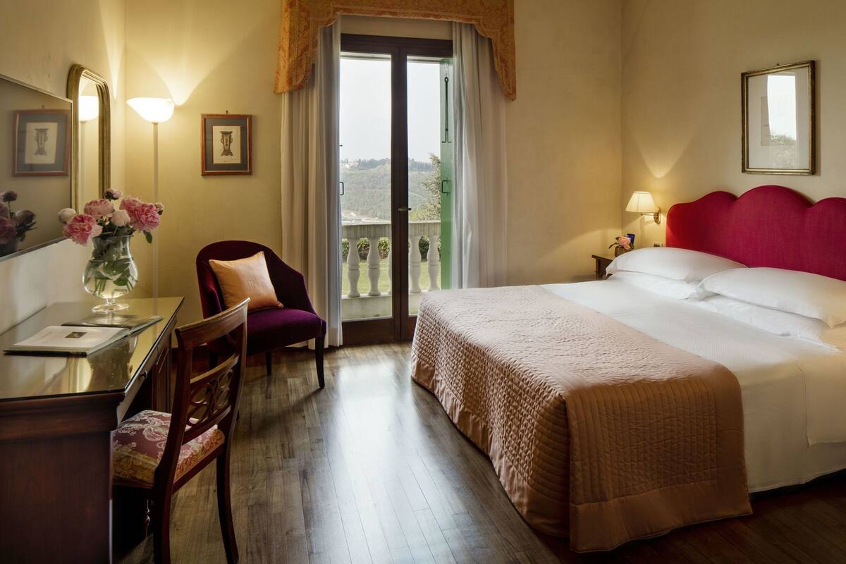 Habitación de hotel con cama, escritorio con silla, un jarrón de flores y balcón con vistas.