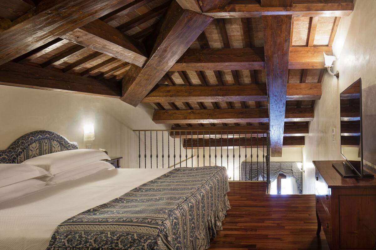 Un dormitorio con techo de madera, una cama con ropa de cama estampada, una mesita de noche y un escritorio con una computadora.