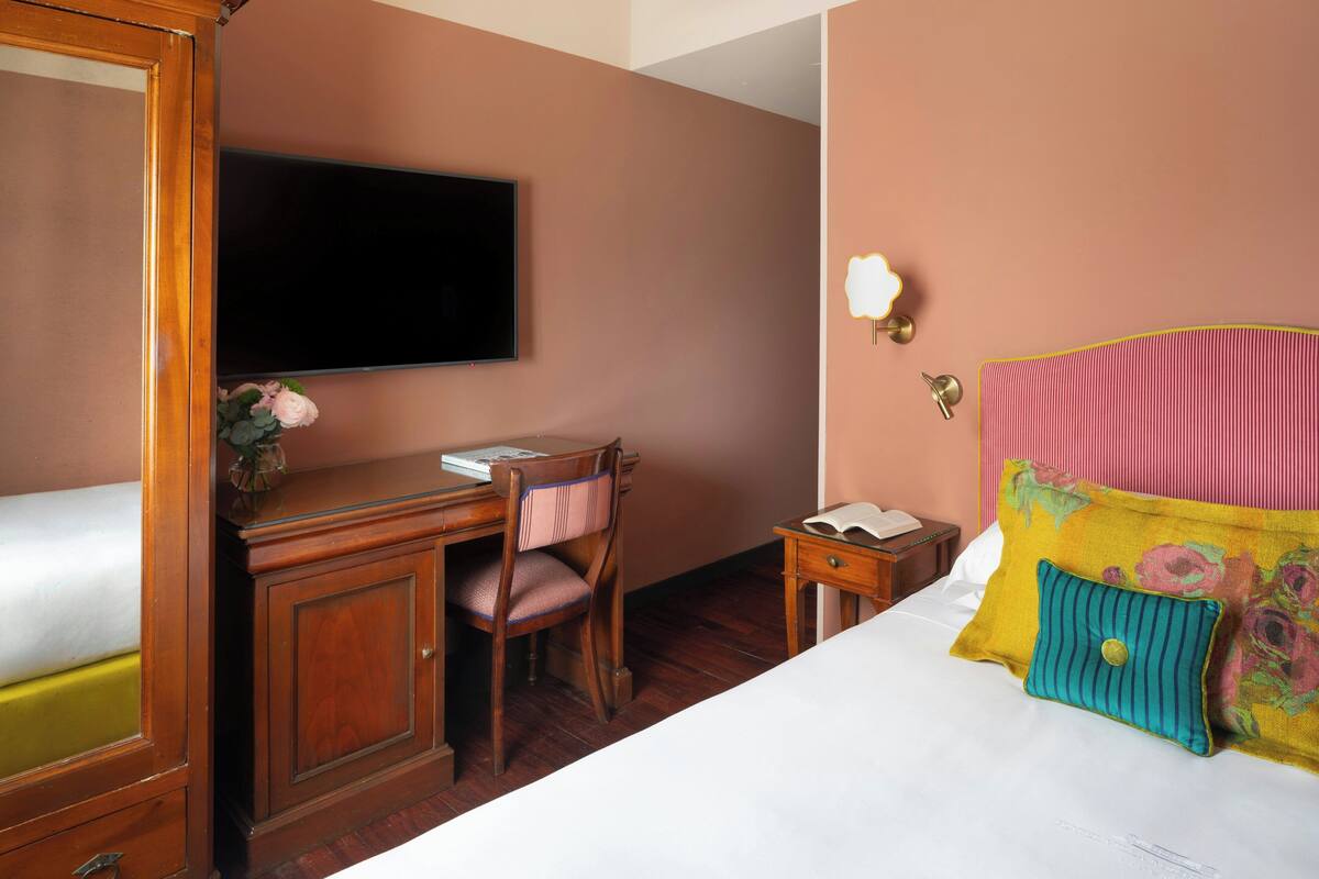 Habitación de hotel con cama, escritorio con silla, televisor, mesita de noche y lámpara fijada a la pared.
