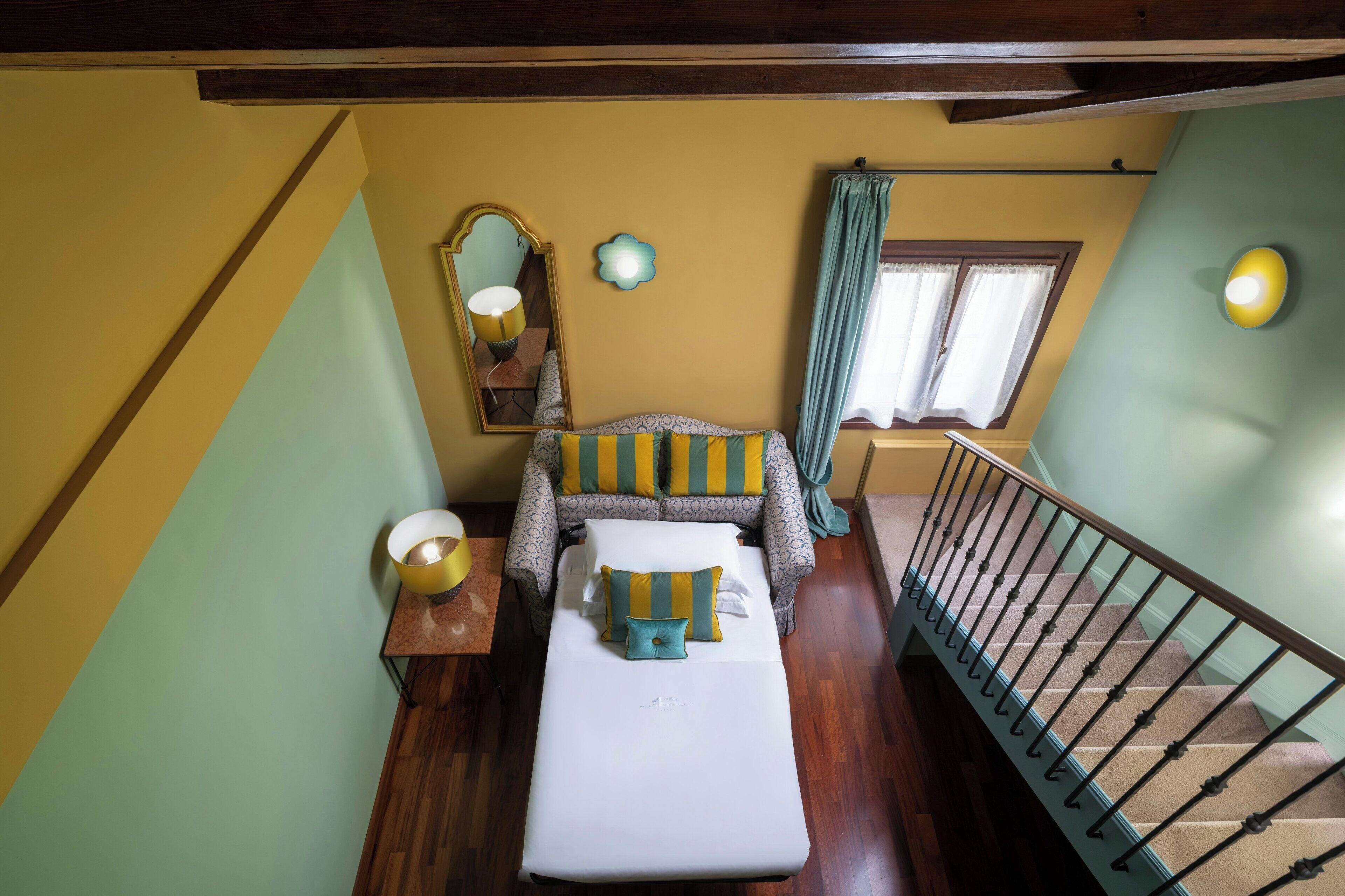Una habitación con una cama, una mesita de noche, un espejo, un ventanal con cortinas y una escalera.