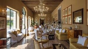 A spacious living room with a high ceiling, large windows, and a chandelier. There are multiple seating areas with sofas and chairs.