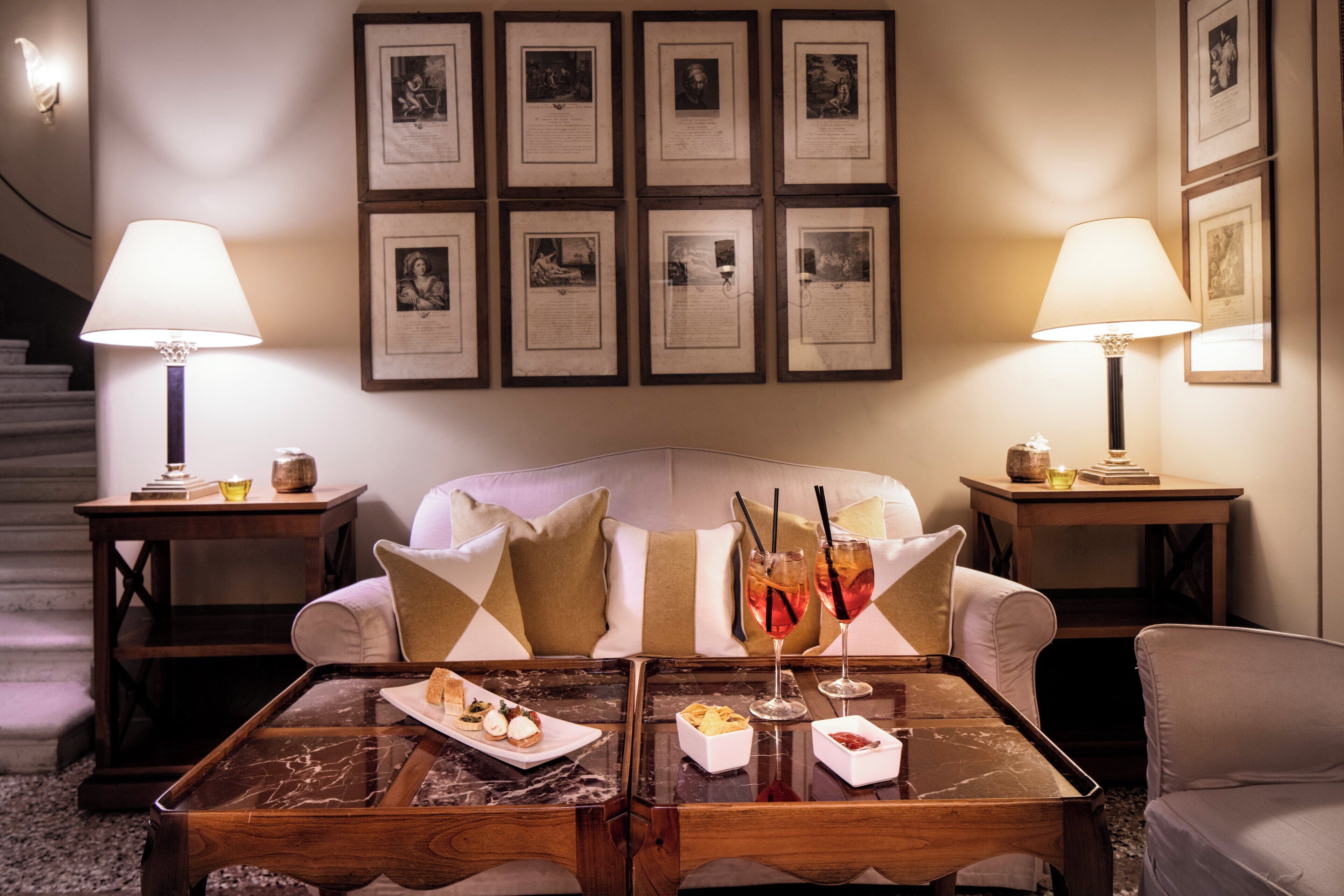 Un rincón acogedor con un sofá, dos mesitas de noche con lámparas, una mesa baja con aperitivos y bebidas, y cuadros enmarcados en la pared.