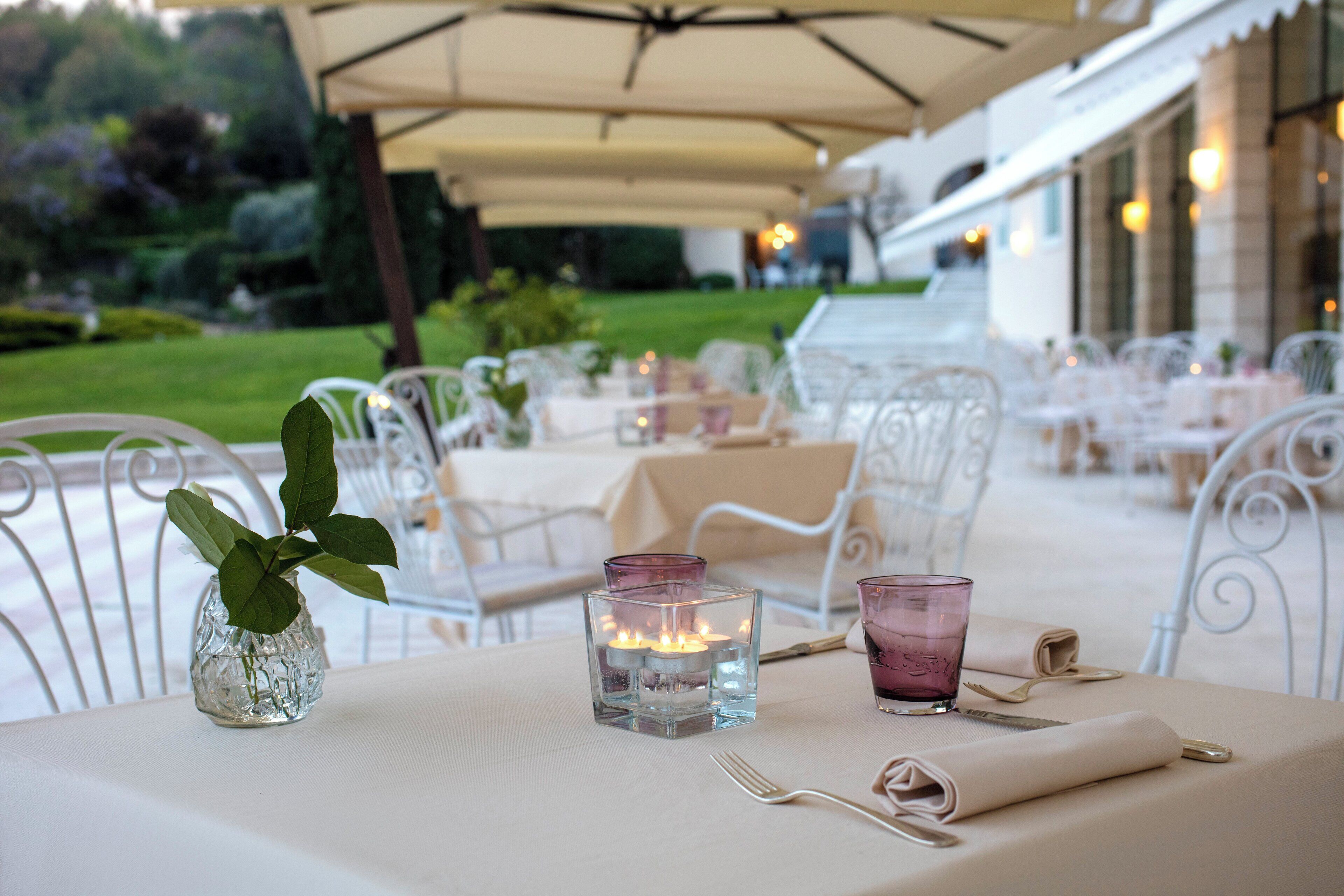 Un área de comedor al aire libre con sillas y mesas de metal blanco, velas y un jarrón con una planta.