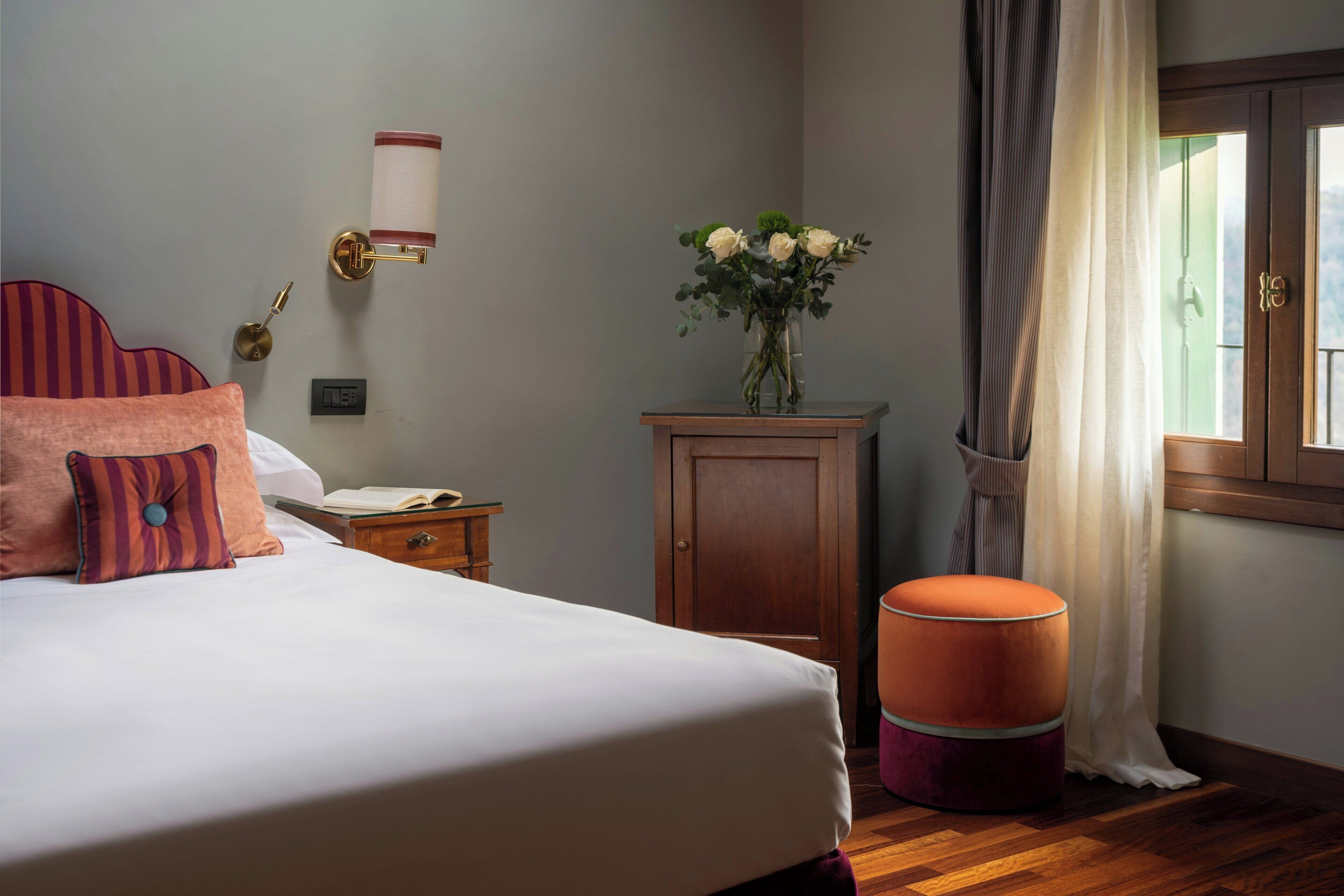 Un dormitorio ordenado con cama, mesita de noche, pequeño armario de madera, florero con flores y ventana con cortinas.