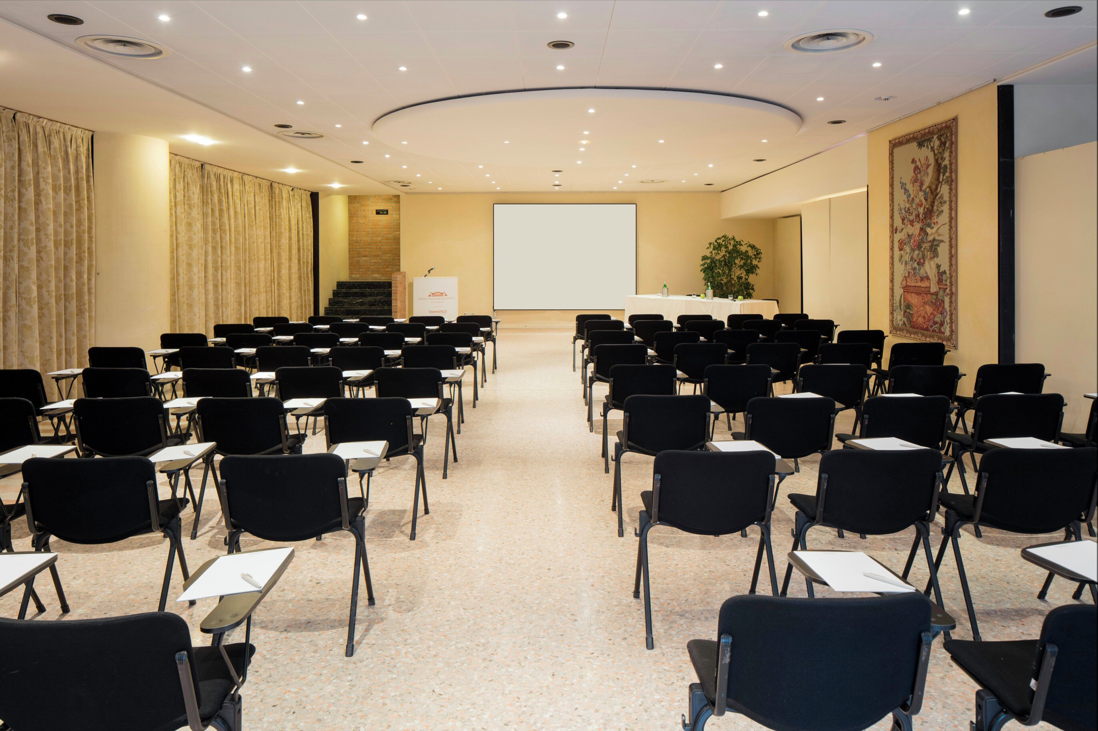 Un salón de conferencias con filas de sillas, una pantalla de proyector, un atril y una escalera.