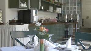 A dining area with a table set for two, featuring a vase with flowers, a candle, and a glass. There are chairs and a shelf with kitchen items in the background.