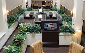 A spacious lobby with multiple seating areas, a central TV displaying a news program, and lush green plants.