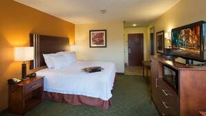 A hotel room with a bed, bedside lamps, a TV, a desk with a phone, and a door leading to another room. - Hampton Inn Cornelia (Cornelia)