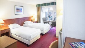A hotel room with two beds, a desk, a chair, a lamp, and a window with a view of buildings. - Delta Hotel City Center (Amsterdam)