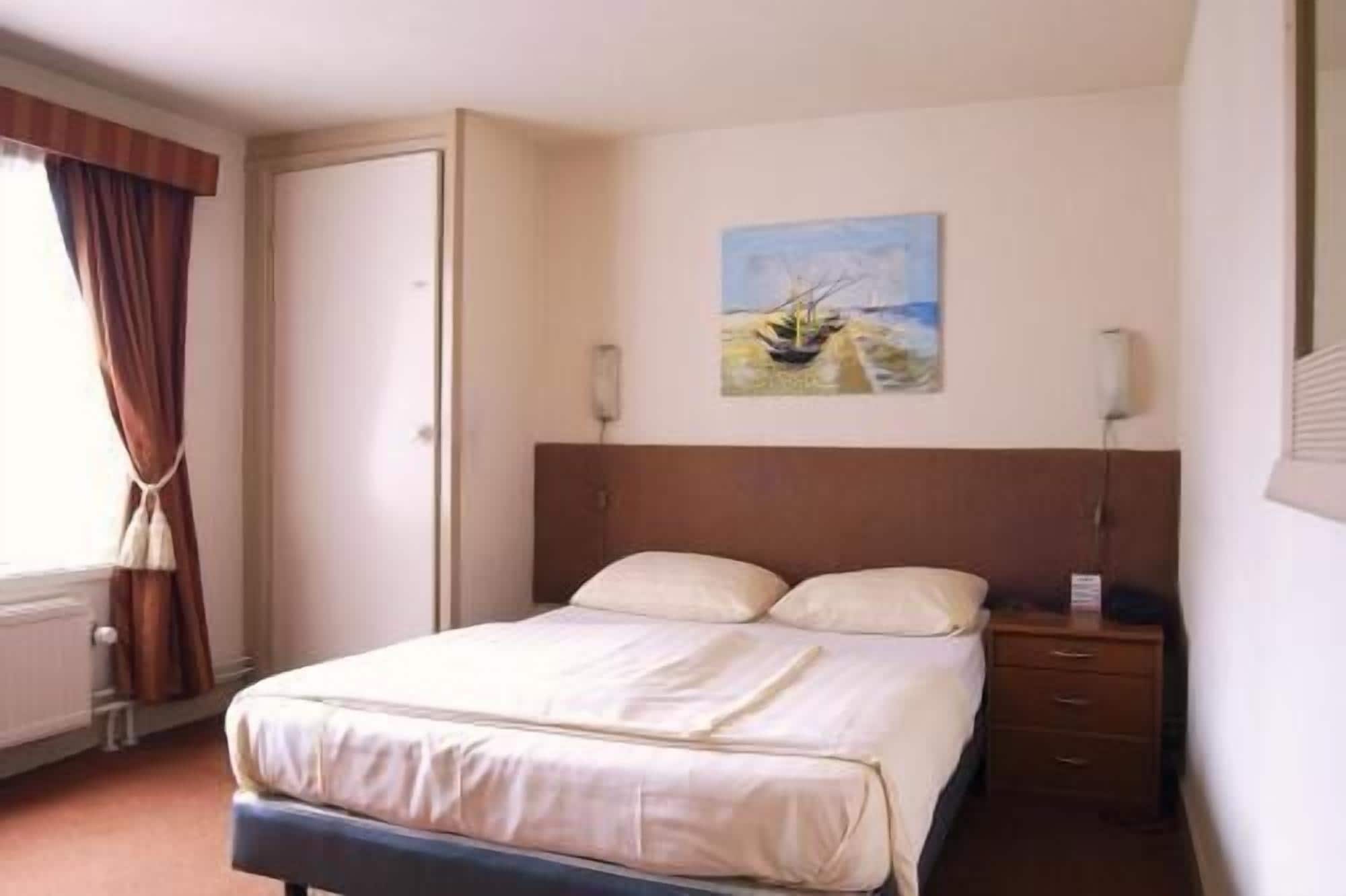 A hotel room with a double bed, a bedside table, a nightstand, a painting on the wall, and a window with curtains.