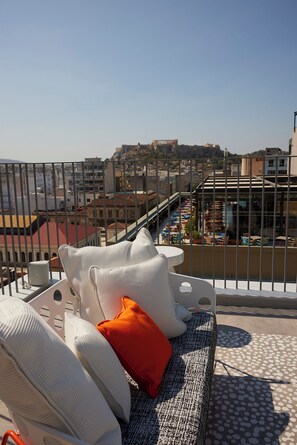 A balcony with a white cushioned seating area, a red pillow, and a view of a cityscape including a hilltop structure.