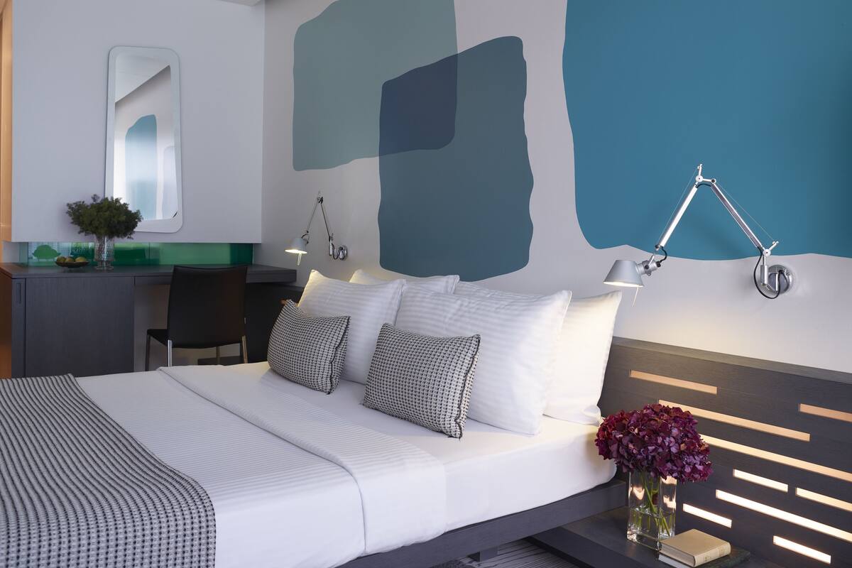 Una habitación de hotel moderna con una cama grande, lámparas de noche, un escritorio con silla y una pared decorada con arte abstracto.