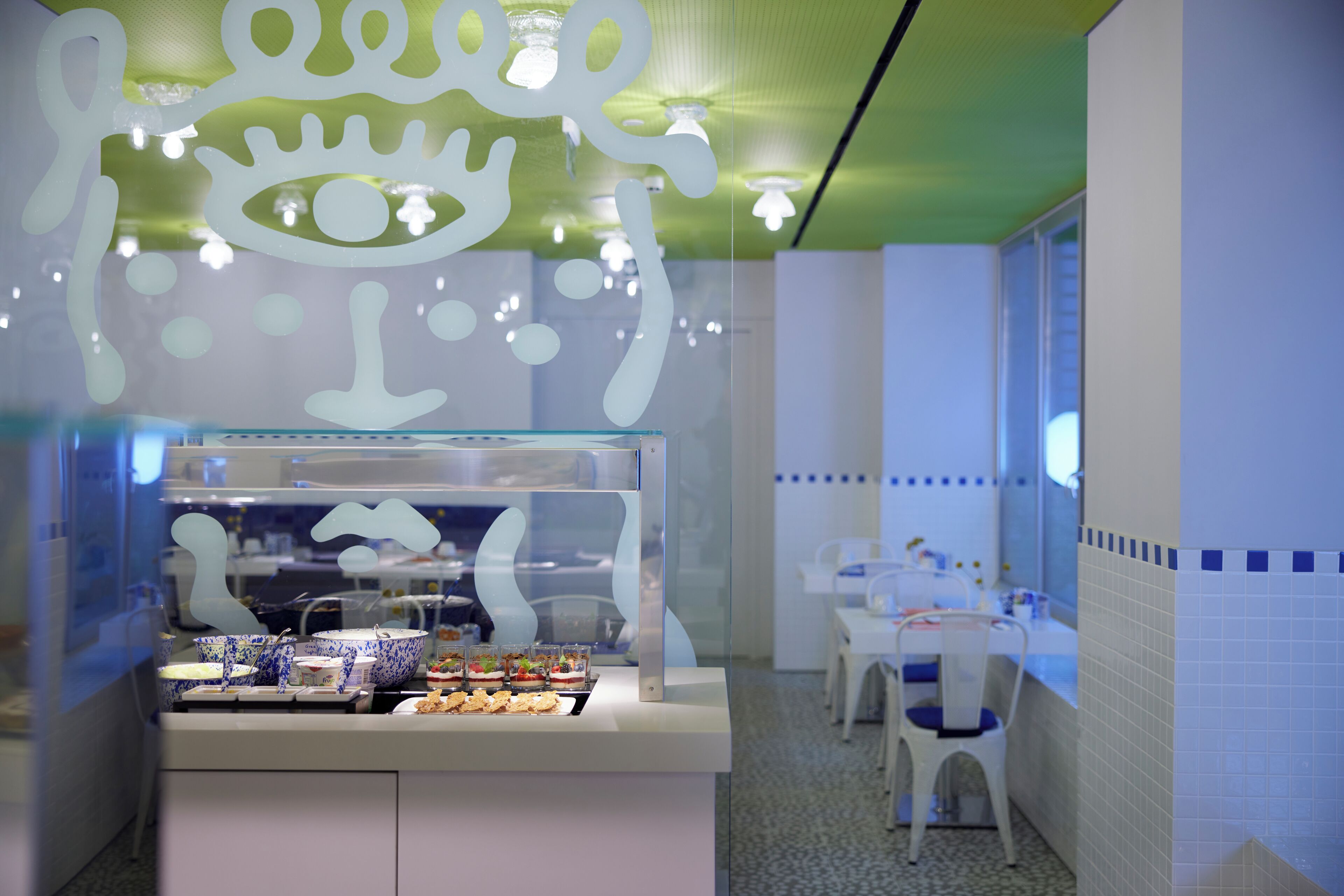 A cafe with a glass display case containing food items, a white counter, and a seating area with tables and chairs.