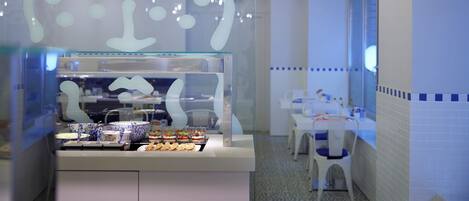 A cafe with a glass display case containing food items, a white counter, and a seating area with tables and chairs.