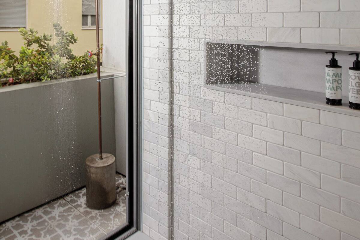 Una ducha moderna con cabezal de lluvia, una ventana con vista a un edificio y un estante pequeño con artículos de tocador.