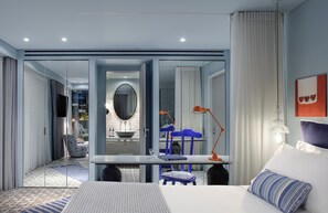 A modern hotel room with a glass partition, a bed, a desk, and a view of the bathroom.