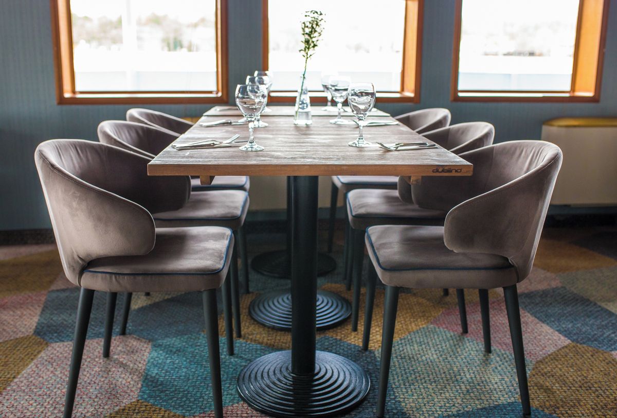 Una mesa de comedor con seis sillas, un florero con planta y copas de vino sobre una alfombra estampada.