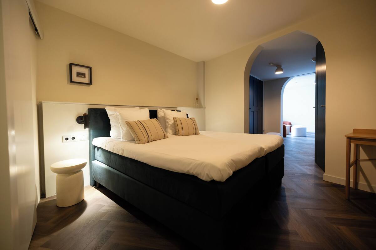 A bedroom with a bed, a bedside table, a toilet, and a hallway leading to another room.
