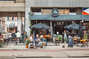 An outdoor cafe with tables and chairs, a sign for Baker & Moore, and a flag. - Hotel Emma (Rotterdam)