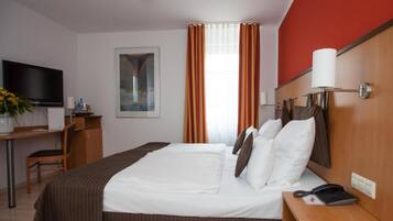 Habitación de hotel con cama, escritorio, televisor, un cuadro y ventana con cortinas.