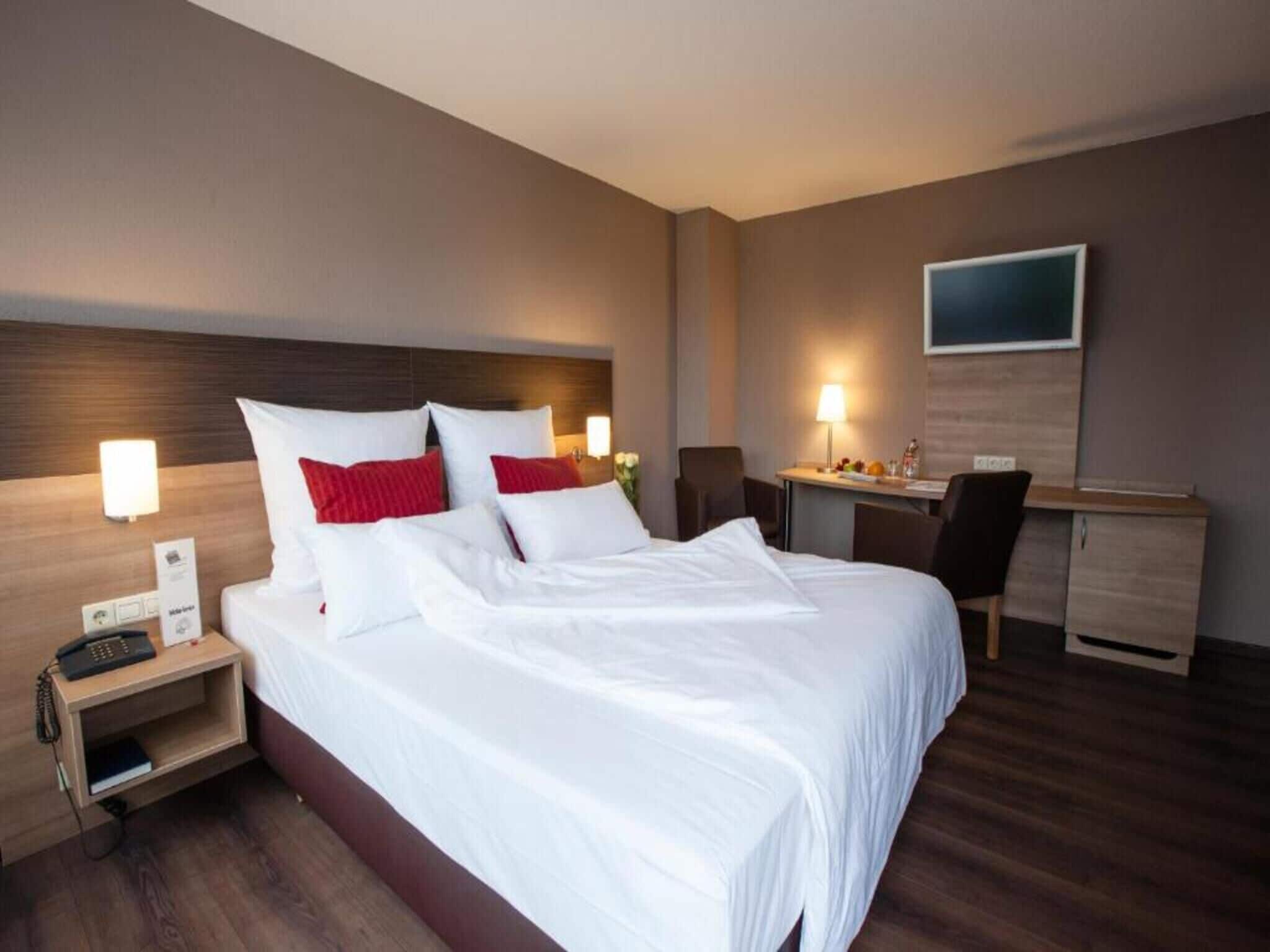 A hotel room with a large bed, bedside table with a phone, a desk with a chair, and a flat-screen TV on the wall.