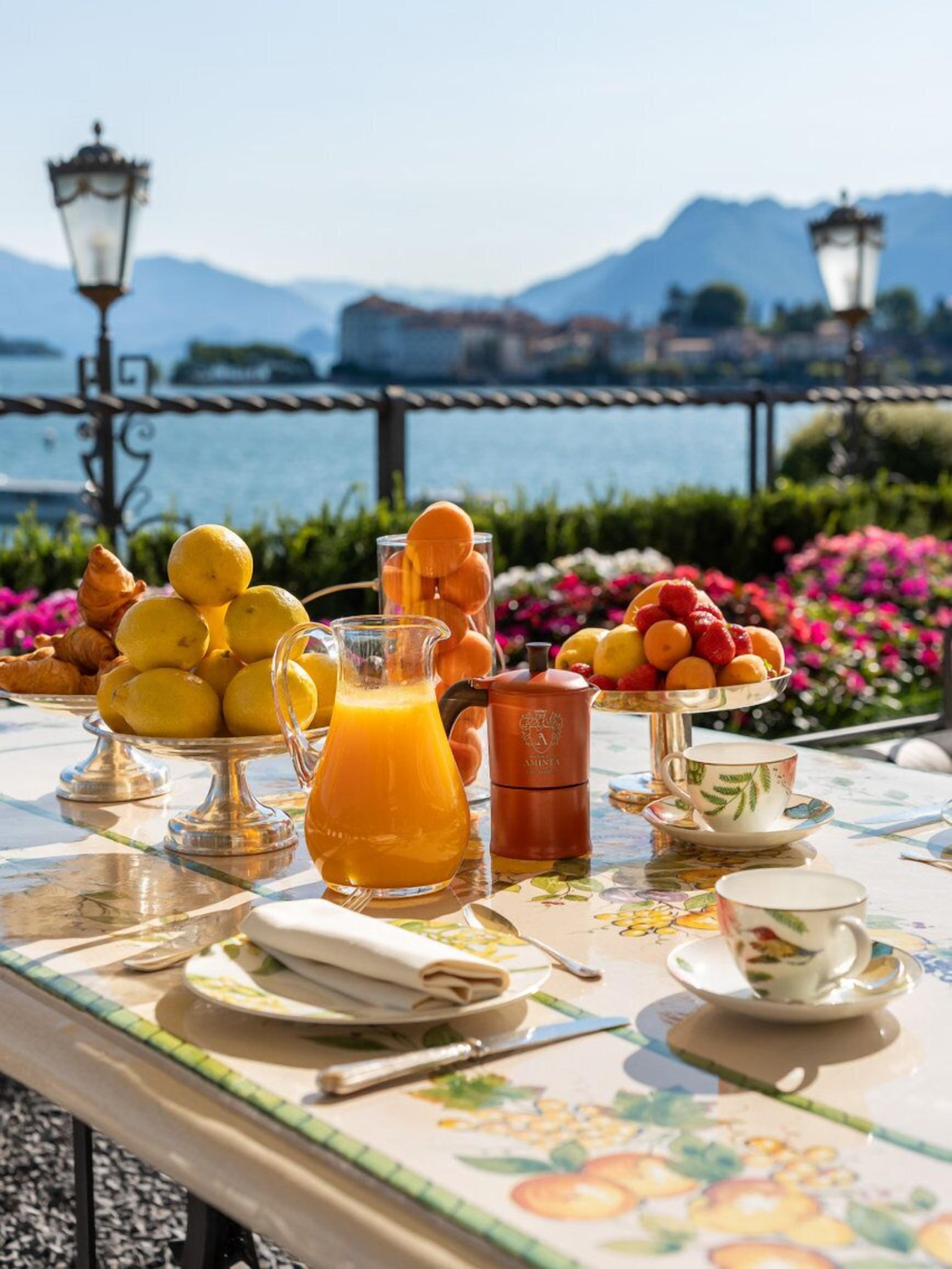 A table set with breakfast items including fruits, pastries, and a jug of orange juice, overlooking a scenic lake with mountains in the background.