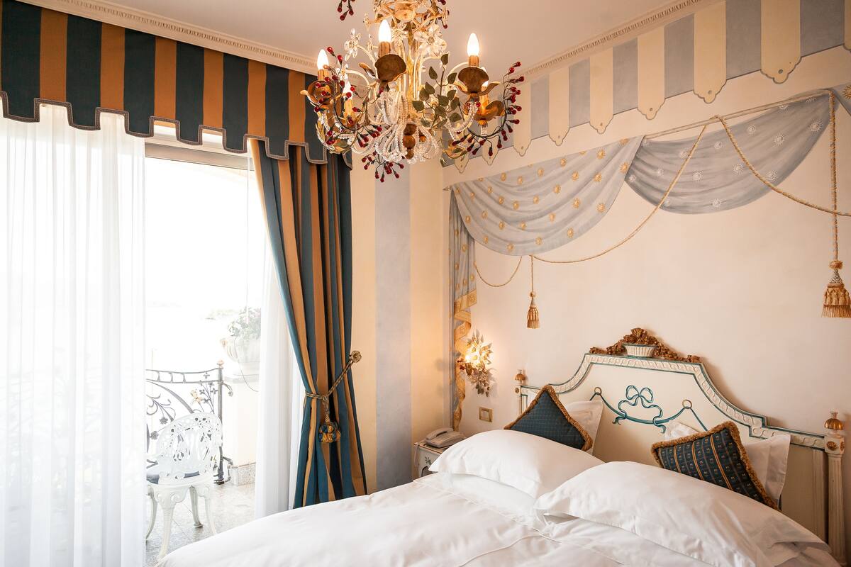 A bedroom with a large bed, a chandelier, curtains, and a view of a balcony.