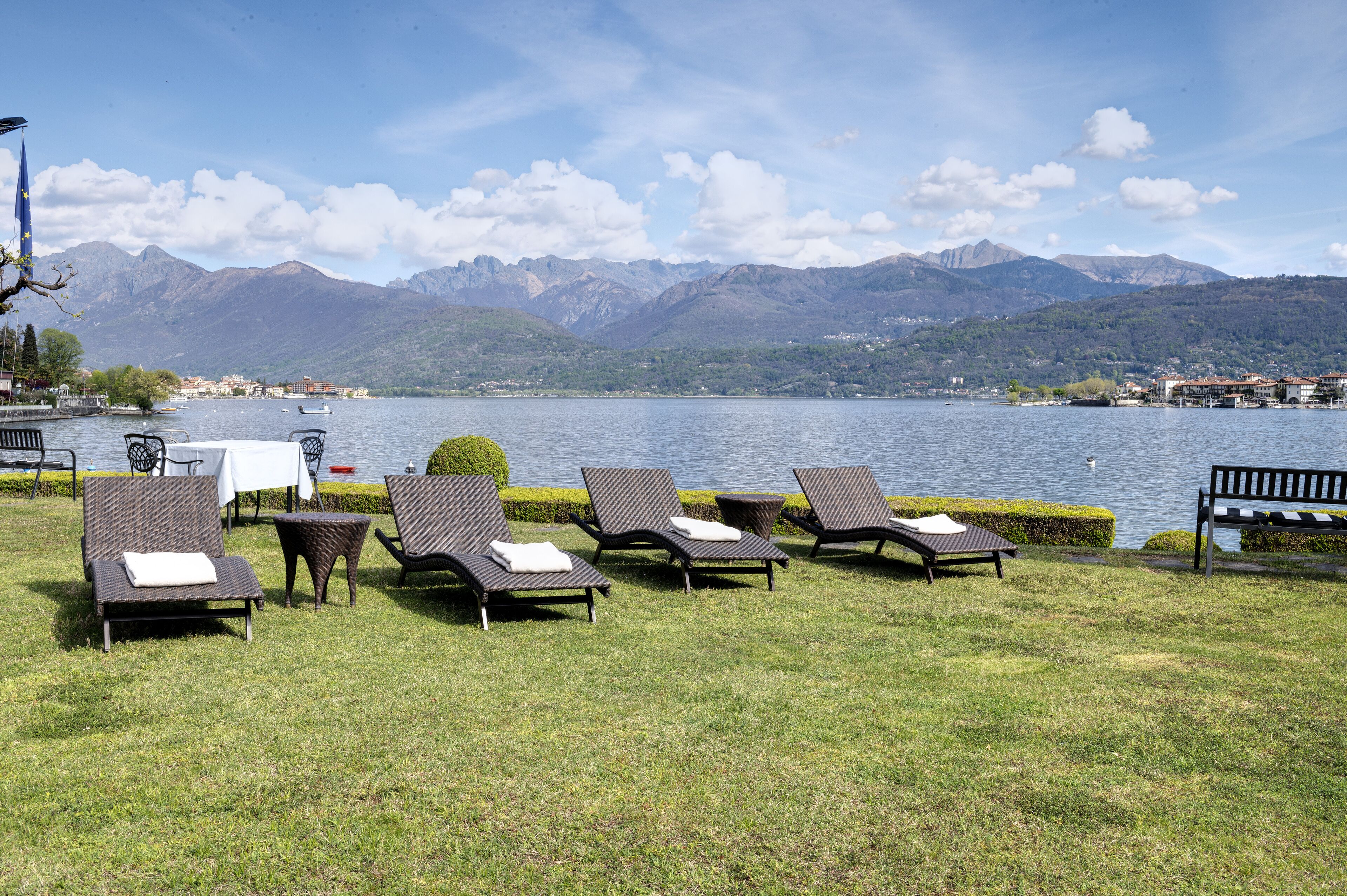 A lakeside lounge area with wicker lounge chairs and a small table, overlooking a lake and mountains.
