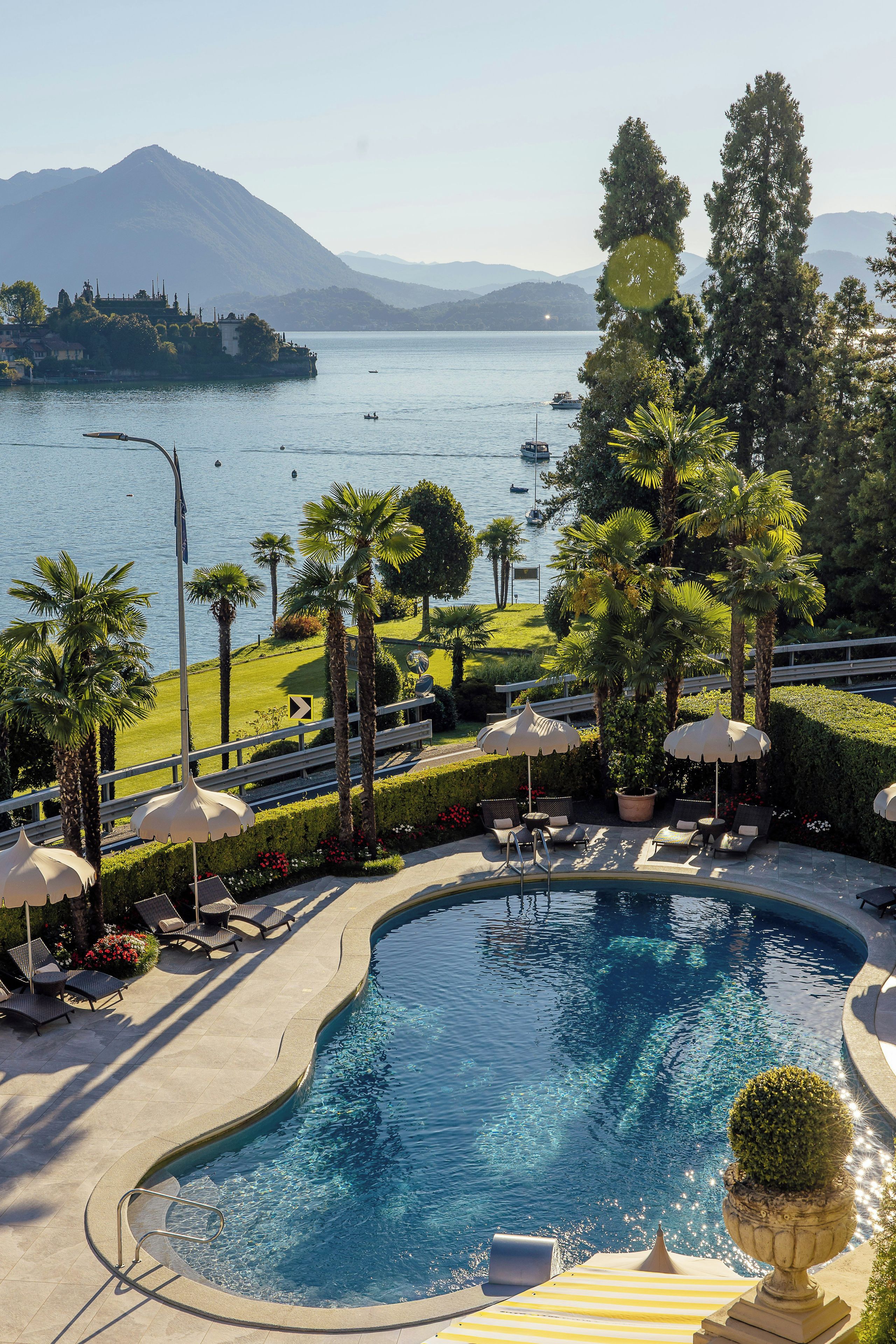 Zona de piscina con sillones de descanso y sombrillas, rodeada de vegetación y con vista a un lago con barcos.