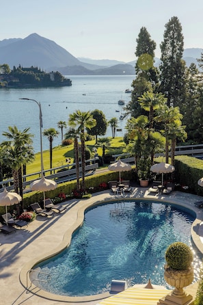 Zona de piscina con sillones de descanso y sombrillas, rodeada de vegetación y con vista a un lago con barcos.