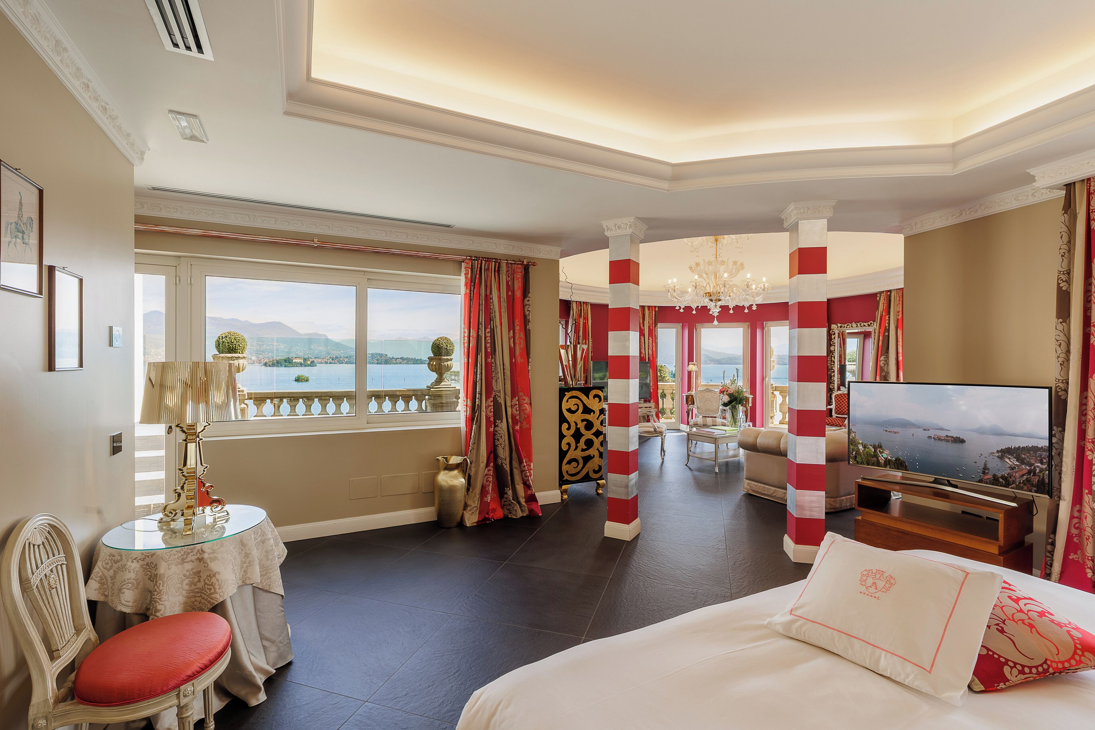 A hotel room with a large bed, a television, a dining table with chairs, and a view of the sea through large windows.
