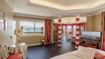 A hotel room with a large bed, a television, a dining table with chairs, and a view of the sea through large windows.