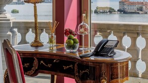 A room with a view of a lake and mountains, a desk with a lamp, a vase with flowers, and a phone.