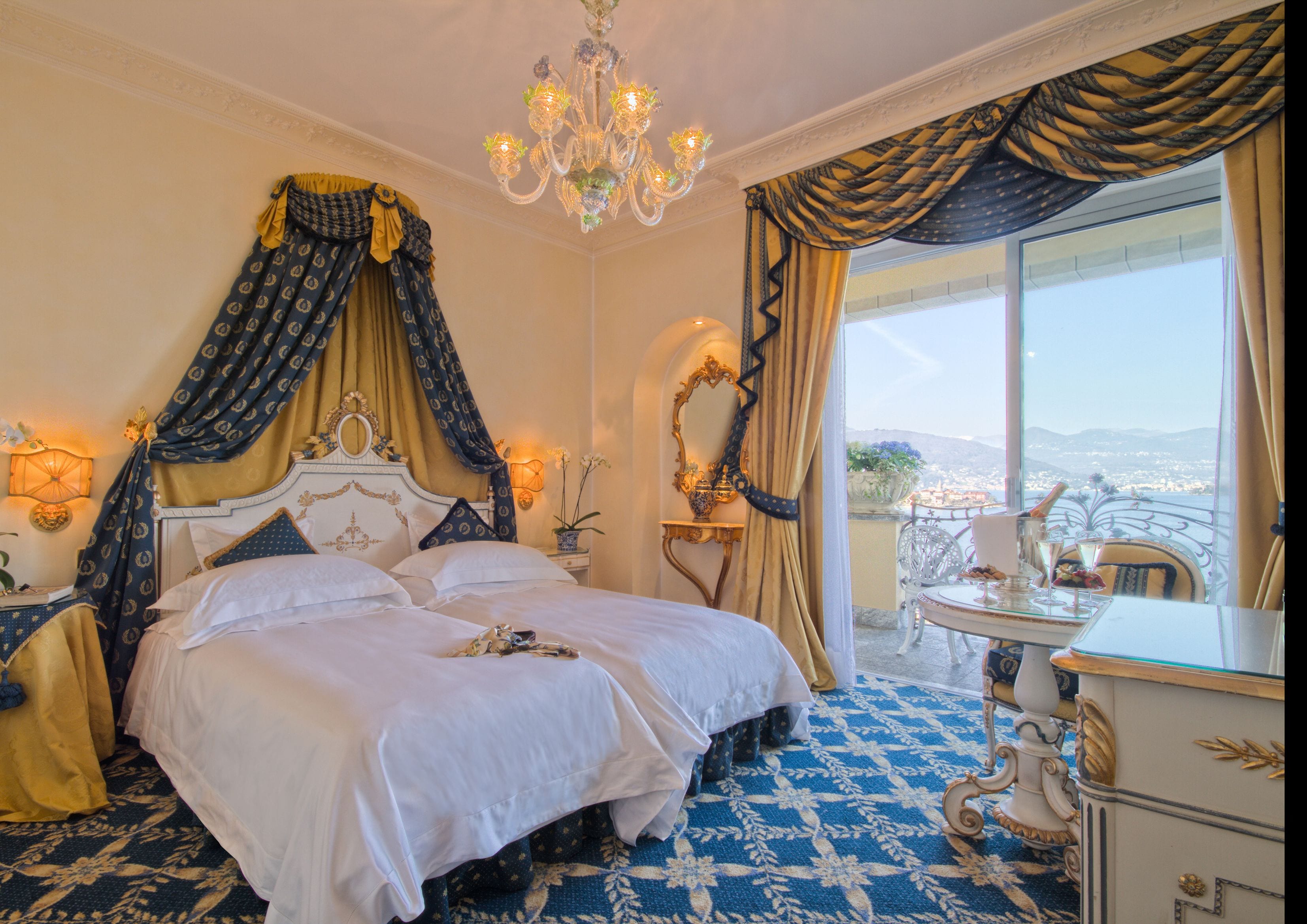 A luxurious bedroom with a large bed, ornate curtains, a chandelier, and a view of the city.