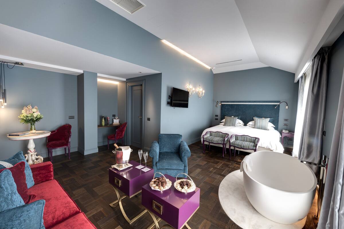 Una habitación de hotel moderna con una cama grande, una zona de estar con un sofá rojo y un sillón azul, una bañera blanca y un televisor de pantalla plana montado en la pared.