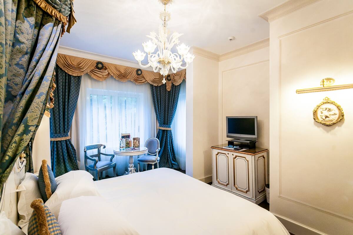 A hotel room with a large bed, a television on a cabinet, a small table with chairs, and a chandelier.