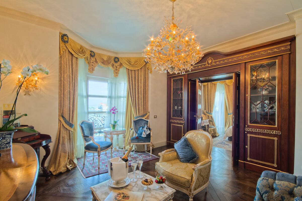 A luxurious room with a chandelier, ornate chairs, a dining table with a bottle of champagne, and a view of the outside through large windows.