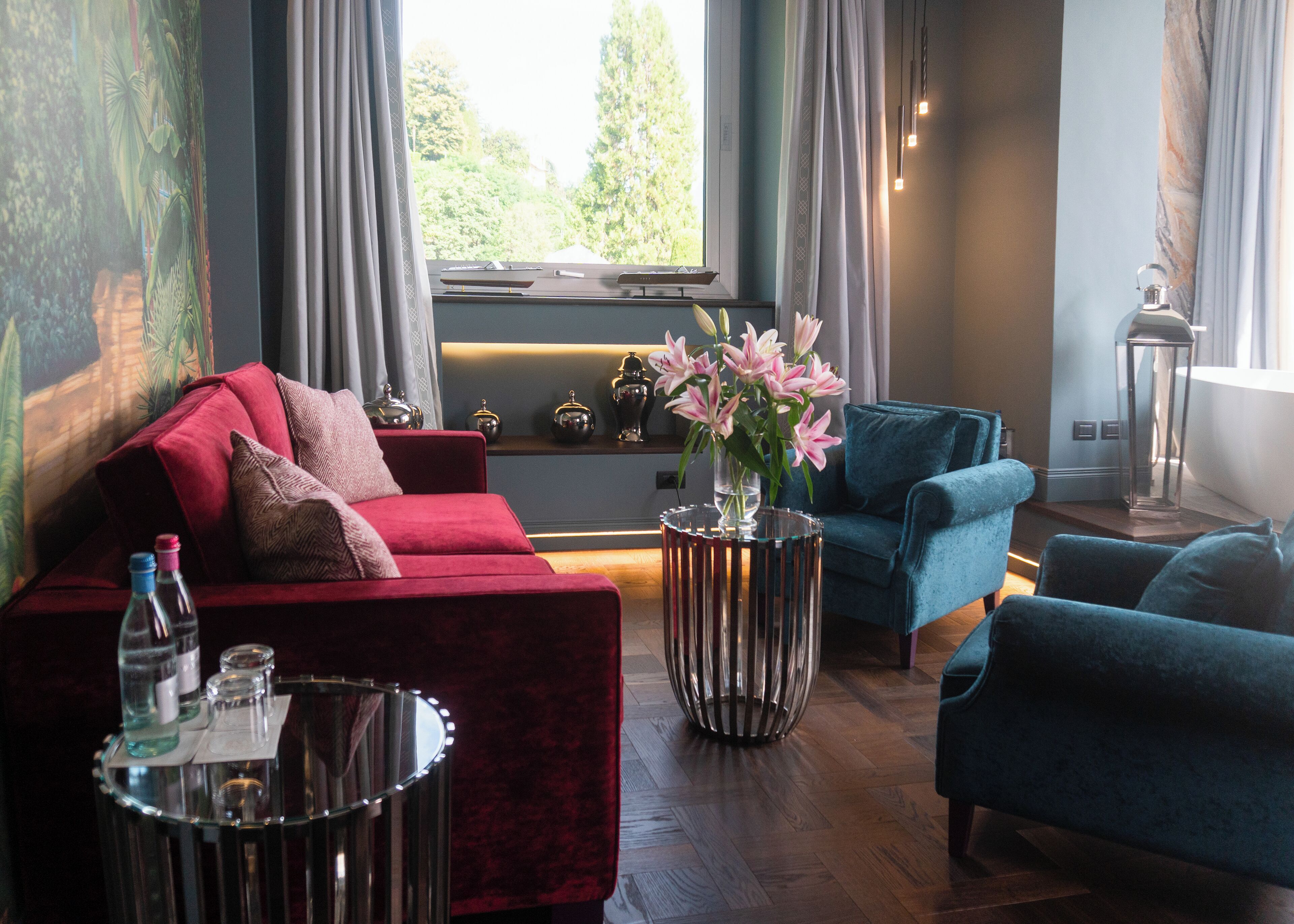 Un salon moderne avec un canapé rouge, un fauteuil bleu et une table basse en verre. On aperçoit un vase rempli de fleurs sur une table d’appoint, ainsi qu’une fenêtre avec des rideaux.