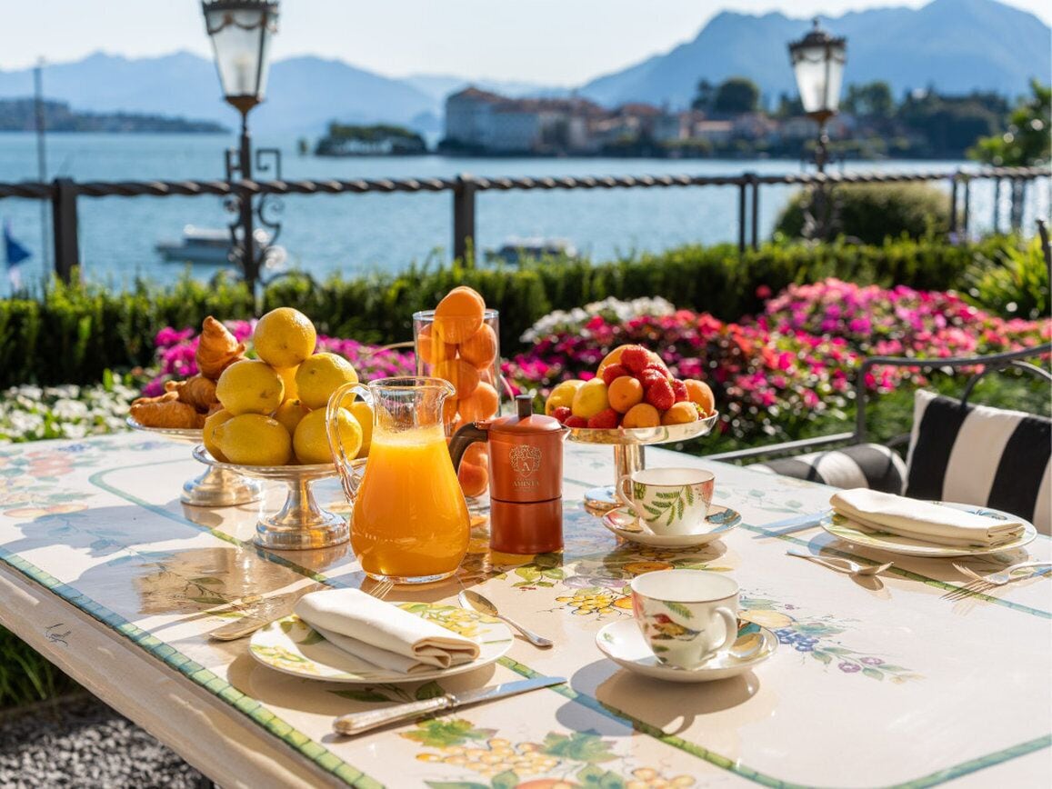 A breakfast table set with fruits, a jug of orange juice, and a coffee maker, overlooking a lake with mountains in the background.