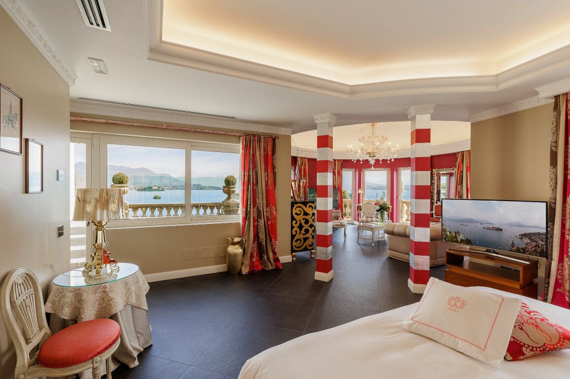 Quarto de hotel com uma cama grande, televisão, área de refeições com mesa e cadeiras, e vista para o mar através de amplas janelas.
