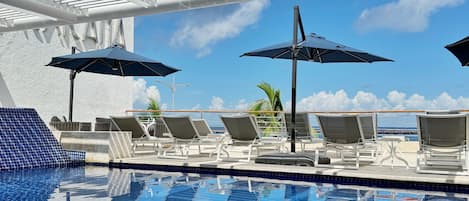Un espace piscine doté de chaises longues et de parasols, offrant une vue dégagée sur l’océan, et une piscine carrelée.