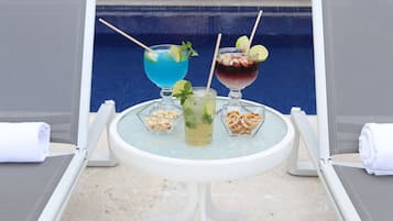 A poolside table with three drinks, each garnished with a lime slice, and two small bowls of nuts.