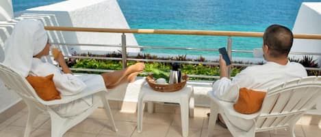 Two white lounge chairs with orange cushions on a balcony overlooking a turquoise sea and a pier.