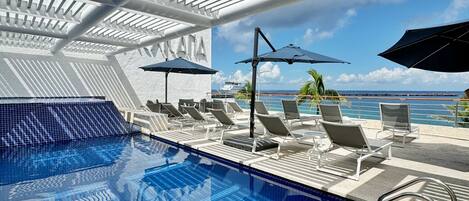 Zona de piscina con sillones de descanso y sombrillas, vista despejada al océano y una piscina con azulejos.