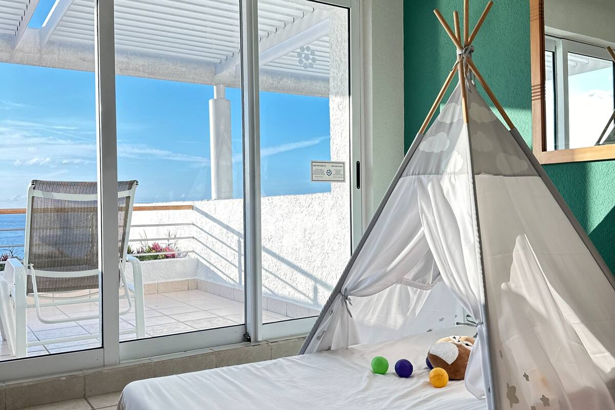 Une chambre avec un lit, une tente indienne blanche et une vue sur la mer par une grande fenêtre.