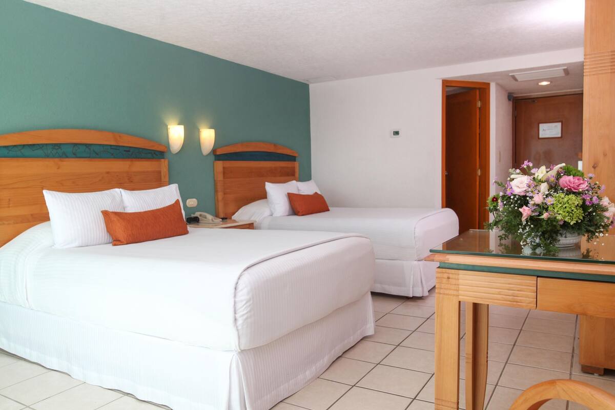 Une chambre d’hôtel avec deux lits, une tête de lit en bois, une petite table sur laquelle est posé un vase de fleurs et une chaise en bois.