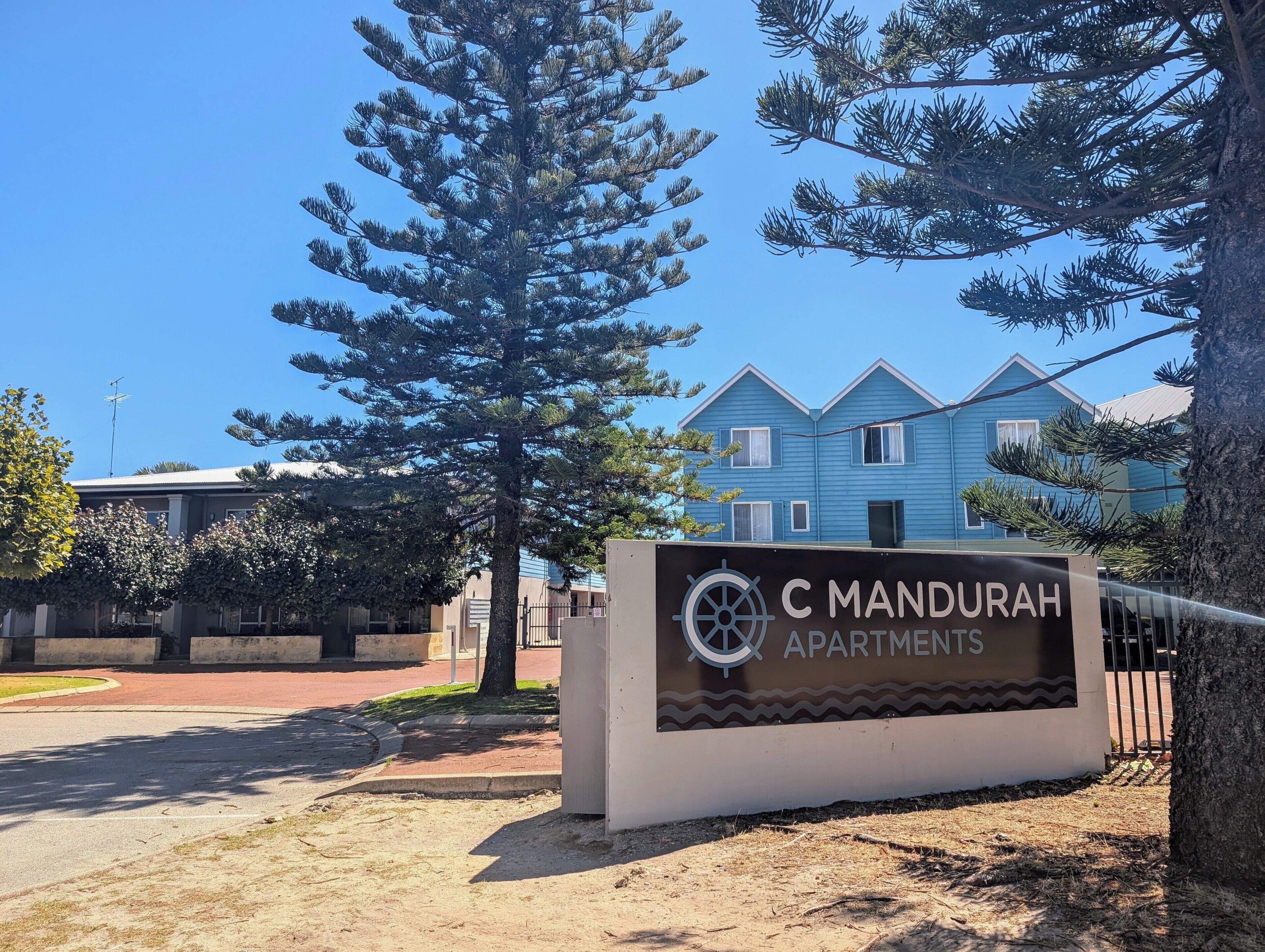 Schild für C Mandurah Apartments, im Hintergrund ist ein klarer Blick auf das Gebäude.