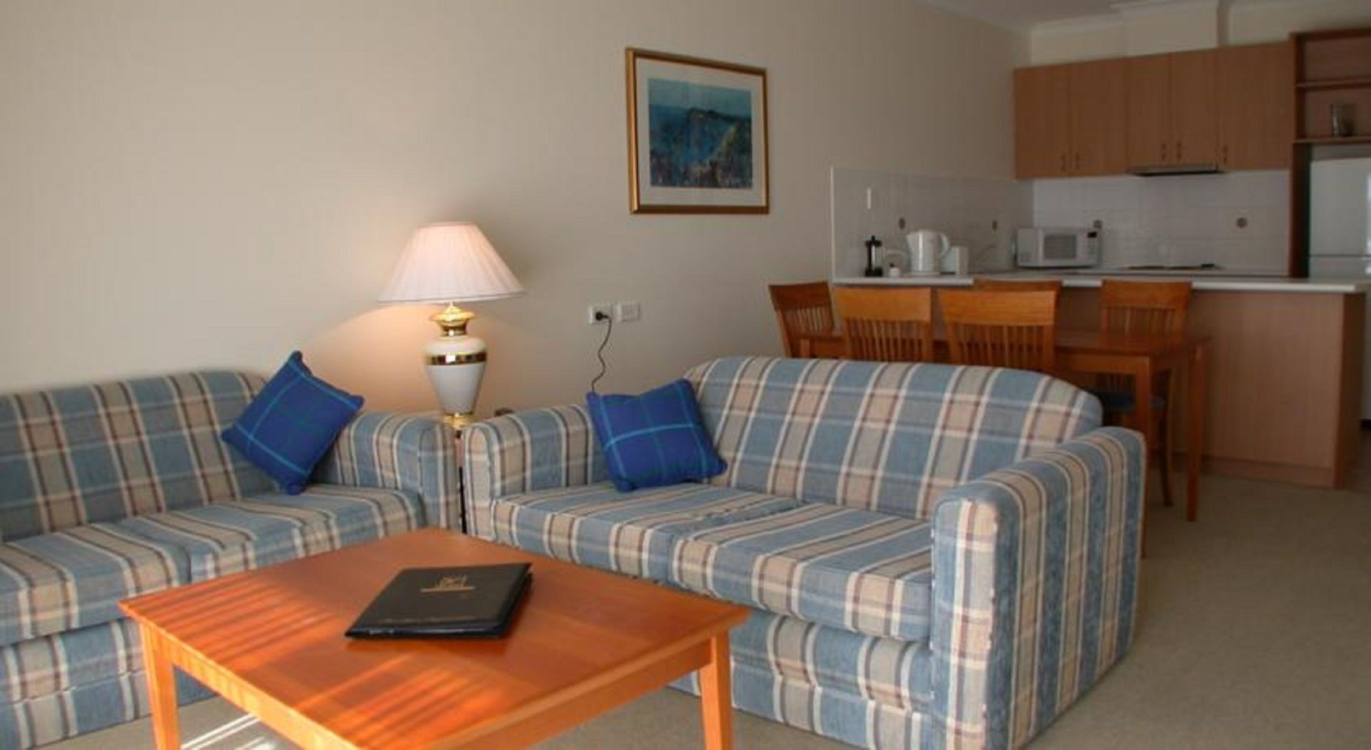 A living room with a sofa set, a coffee table, and a lamp. There is a kitchen area with a microwave and cabinets.