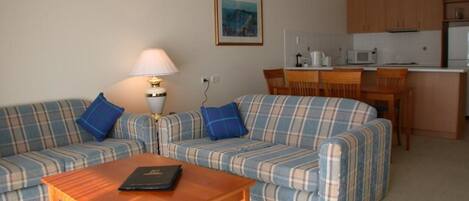 A living room with a sofa set, a coffee table, and a lamp. There is a kitchen area with a microwave and cabinets.